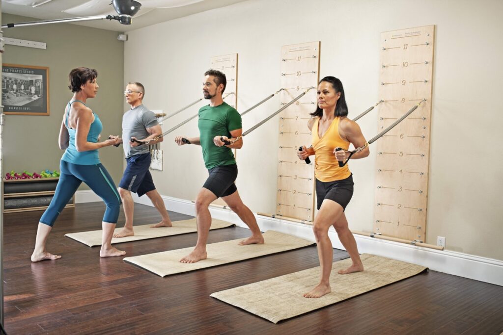Četri cilvēki vingro ar pie sienas piestiprinātām Pilates atsperu iekārtām.