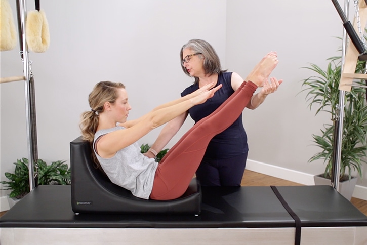 Instruktors palīdz veikt Pilates vingrinājumu uz C-Shaper aprīkojuma.