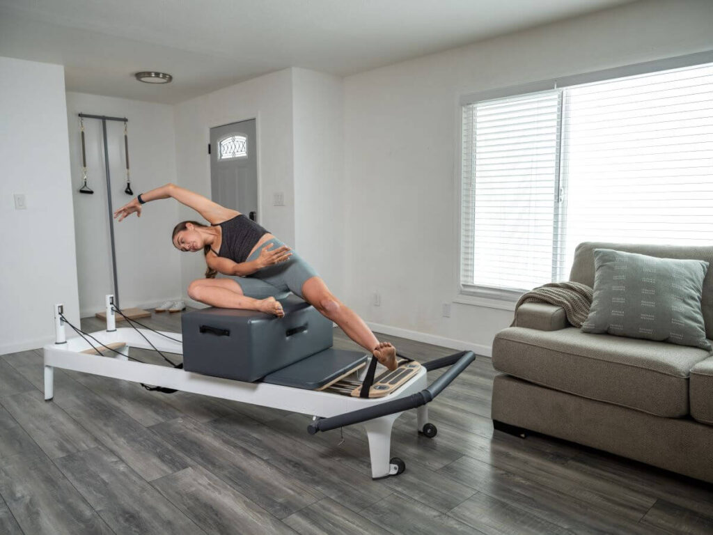 Женщина использует ножные ремни в позе «Русалка» на реформере с сиденьем