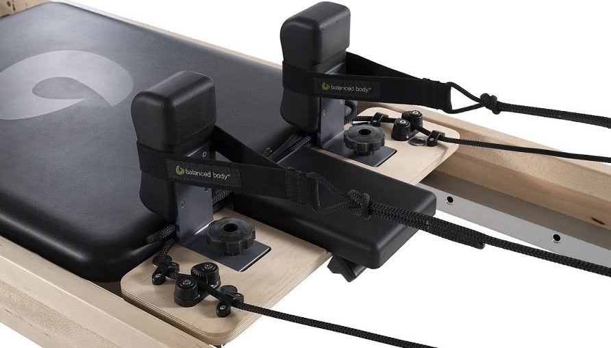 Pilates Reformer kustīgā platforma ar regulējamiem plecu balstiem un melnu polsterējumu.