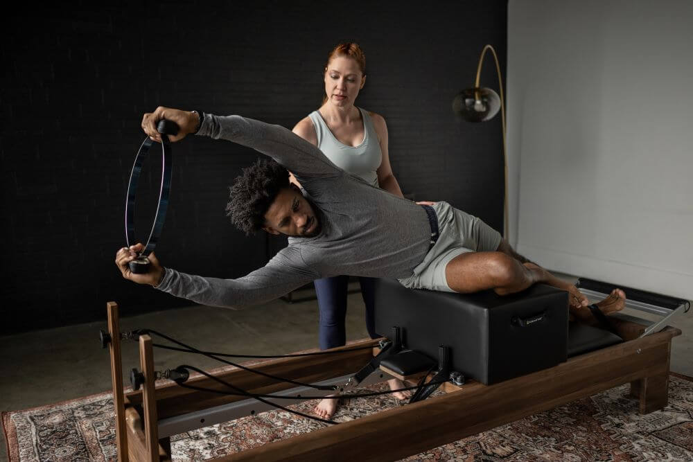 Pilates instruktors palīdz klientam uz standarta sēdēšanas kastes ar reformera iekārtu.